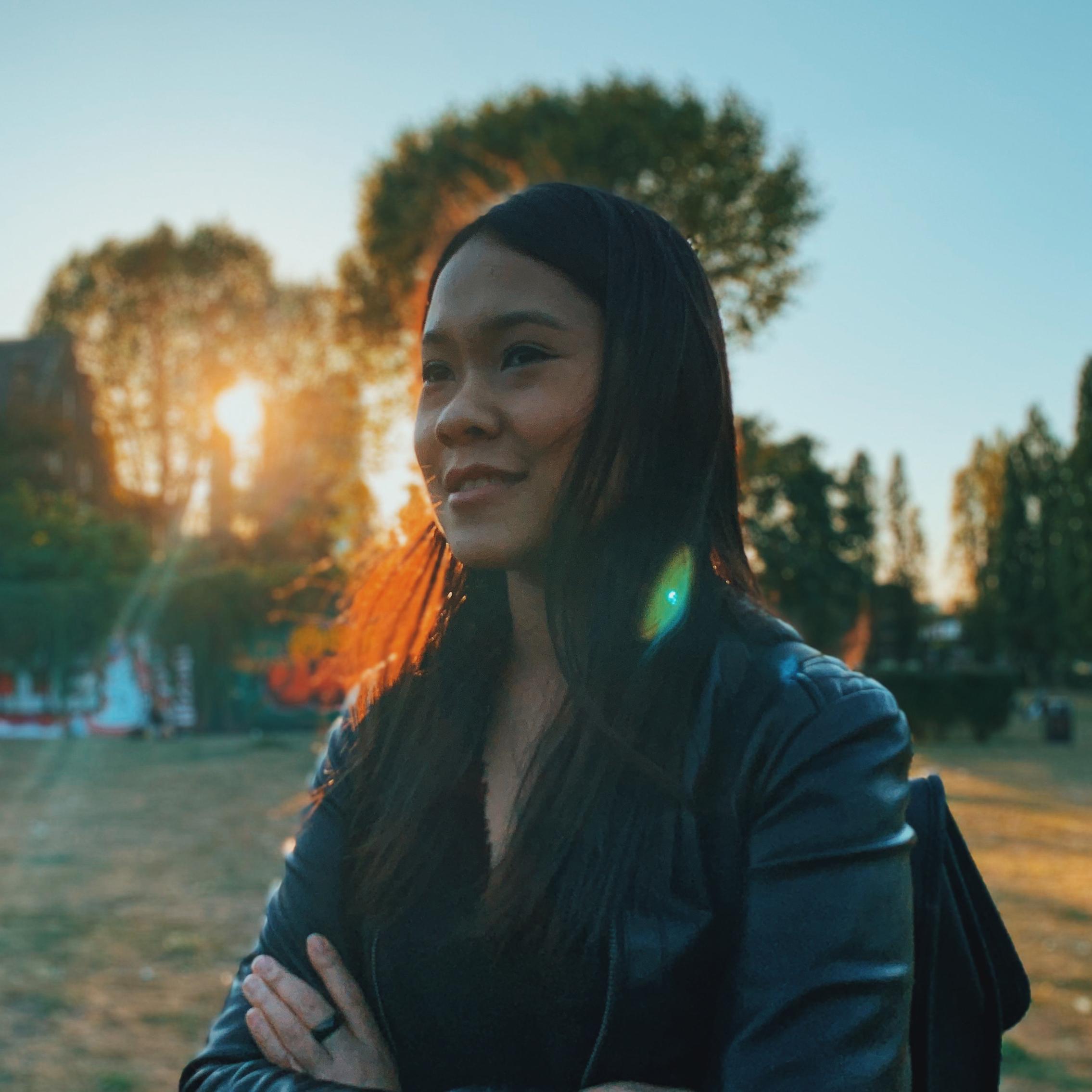 Syaheedah Iskandar, a brown-skinned person with black long hair is standing in front of a park outside looking to the left-hand side of the image. Framed from the waist up, Iskandar is wearing a black leather jacket.