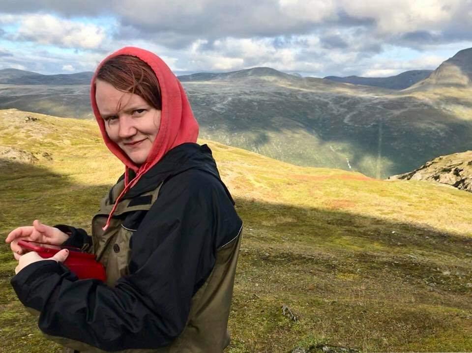 Camilla Fagerli, a white-skinned person with red hair is standing in front of a mountain landscape looking into the camera. Framed from the waist up, Camilla is wearing a black and green outdoor jacket and a red hoodie.