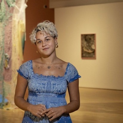Daría Sól Andrews, a brown-skinned person with blonde short hair is standing in front of an exhibition space looking into the camera. Framed from the waist up, Daría is wearing a blue dress and earrings.