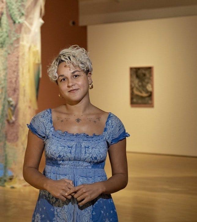 Daría Sól Andrews, a brown-skinned person with blonde short hair is standing in front of an exhibition space looking into the camera. Framed from the waist up, Daría is wearing a blue dress and earrings.