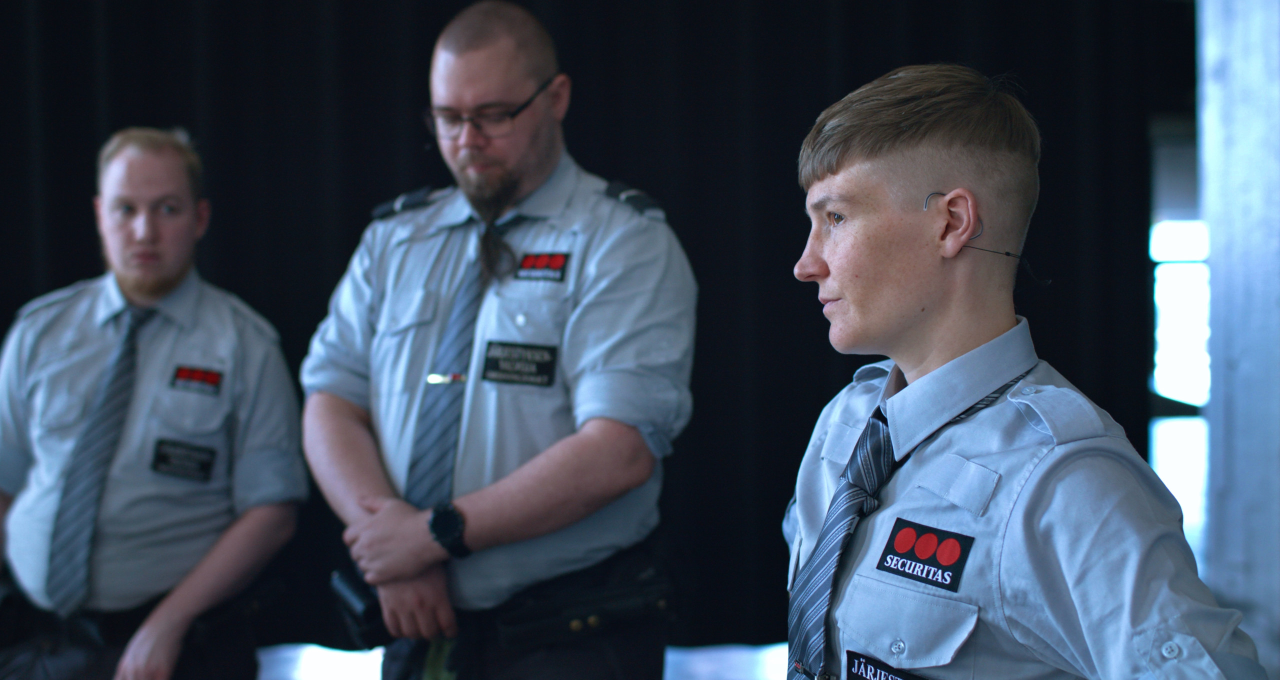 A close-up of three Caucasian guards who seem to be listening to something serious. The guard standing closest to the viewer is the artist Pilvi Takala, and she is looking towards the left of the frame.