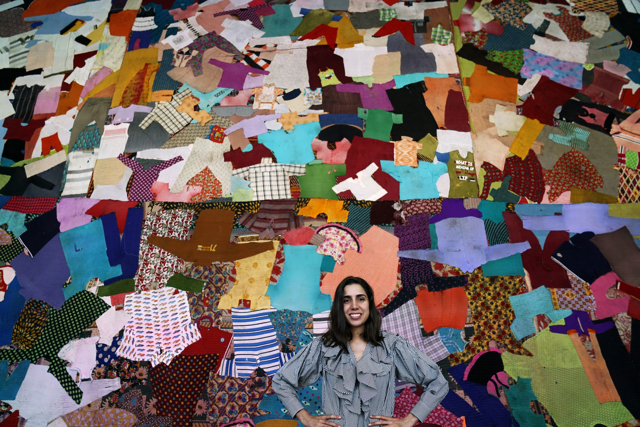 Diana Campbell Betancourt, a light brown-skinned person with shoulder-length brown hair is standing in front of a colourful backdrop comprising of a patchwork of textiles and shirts. Framed from the waist up, Diana is wearing a shirt with white and green stripes. Diana is looking into the camera and smiling while holding her hands on her waist.
