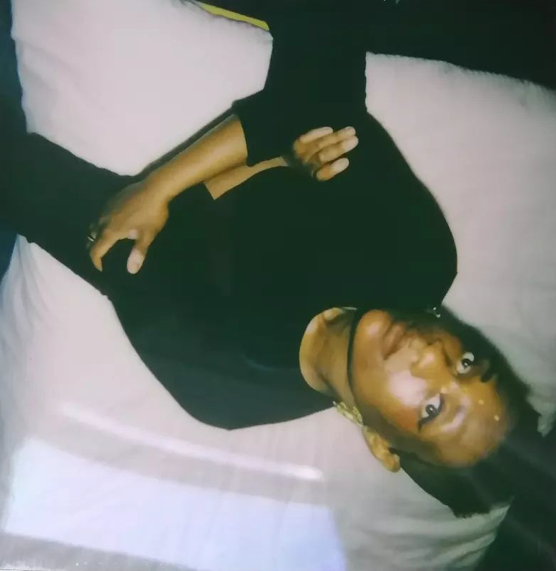An upside-down polaroid image of a black person, wearing black clothes and sitting on a white cushion.