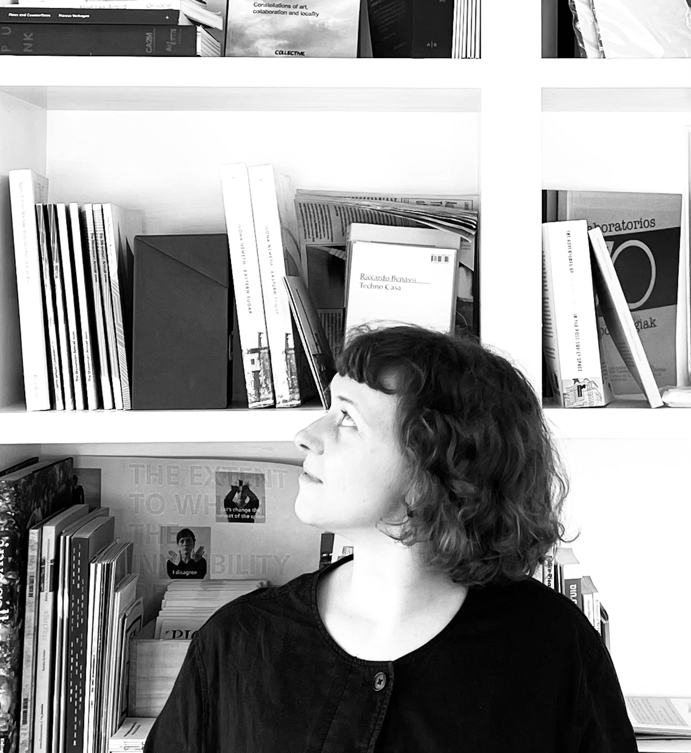 Lily Hall, a white-skinned person with shoulder length curly hair is standing in front of a bookshelf looking in the upright corner of the image. Framed from the chest up, Lily is wearing a dark sweater. The photograph is black and white.