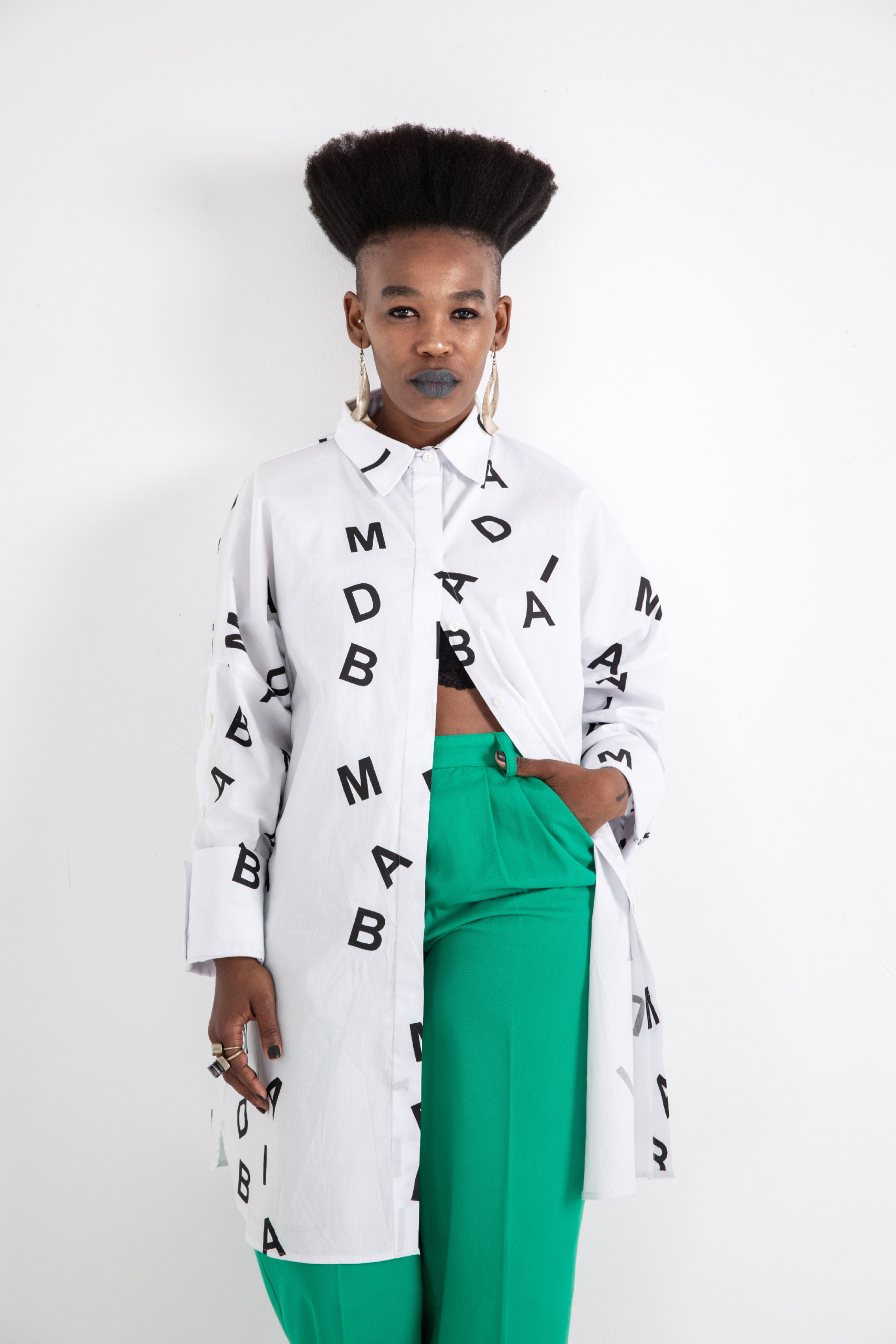 Curator Khansiliye Mbongwa, a blck person with upright curly hair looks straight into the camera, wearing bright green pants and a white coat with typography on it. The words on her coat say MAD.