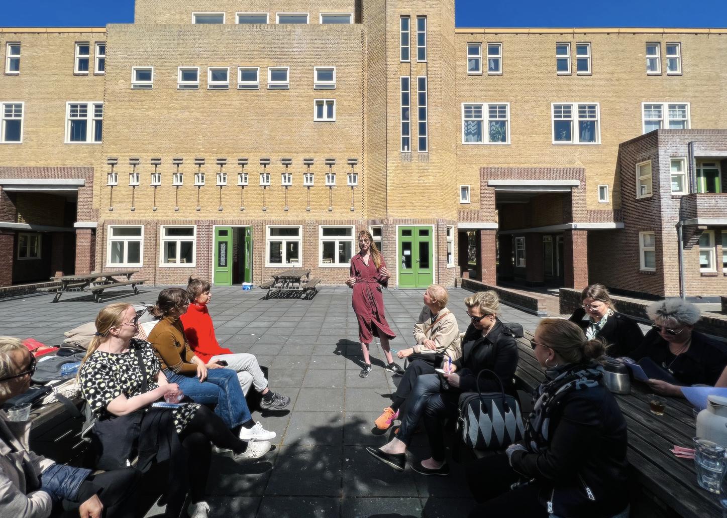 Kuraattori Julia Geerlings seisoo tiilirakennuksen edessä ja ryhmä ihmisiä kuuntelee häntä puhumassa aurinkoisessa säässä.