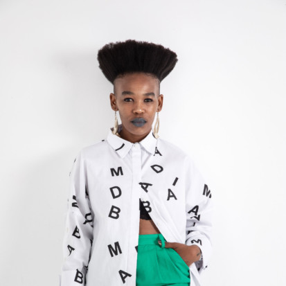 Curator Khansiliye Mbongwa, a blck person with upright curly hair looks straight into the camera, wearing bright green pants and a white coat with typography on it. The words on her coat say MAD.