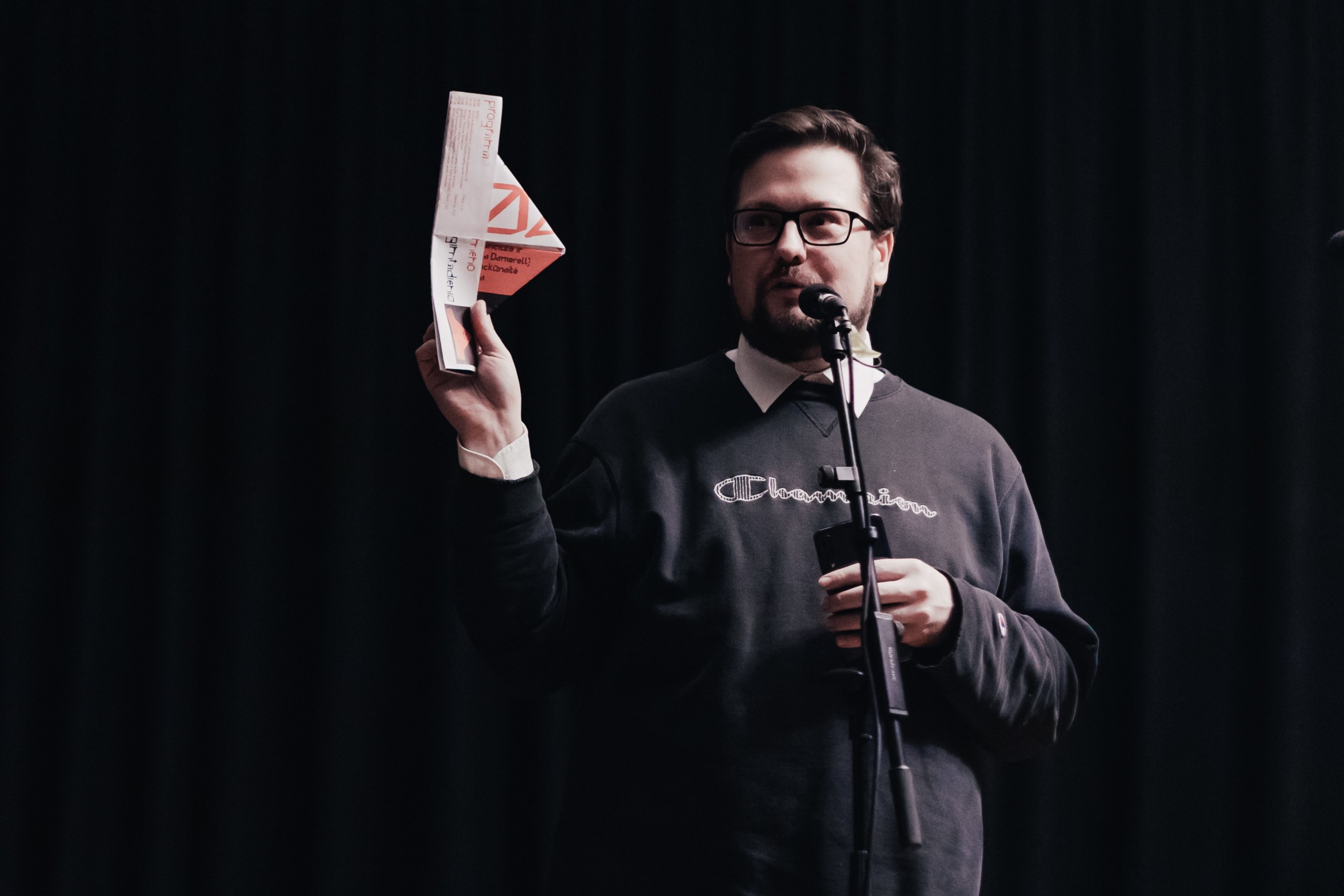 An image of Edvinas Grinkevičius speaking to a microphone and holding a paper hat in his hand.