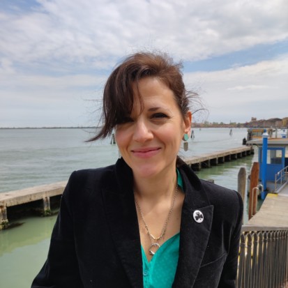 An image of Sara Greavu smiling and looking into the camera, with wind blowing into her hair. In the background is the sea and some sort of a pier.