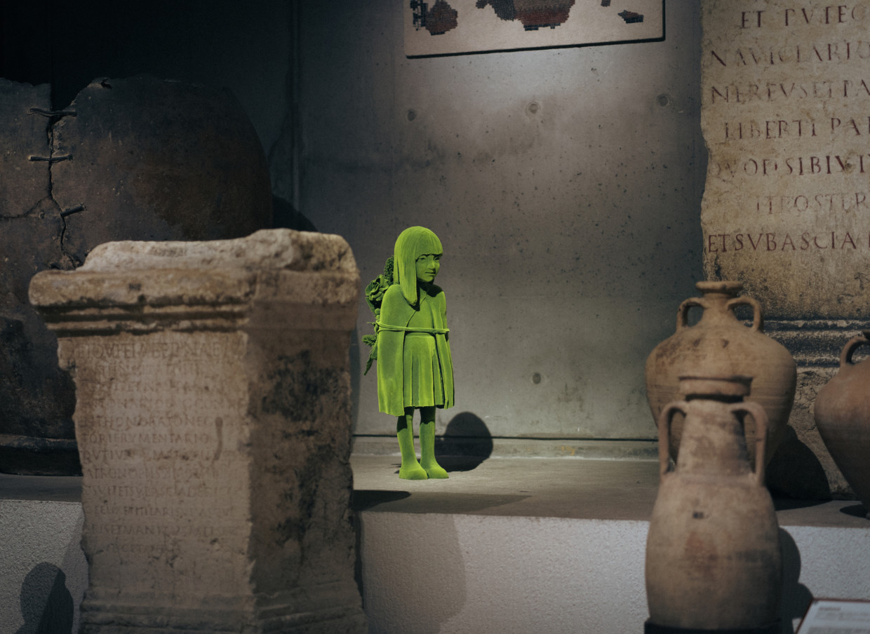 A girl-shaped sculpture, covered in green, moss-like material, standing amongst ancient pots and pillars.