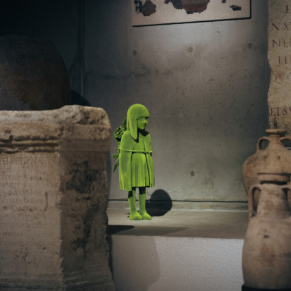 A girl-shaped sculpture, covered in green, moss-like material, standing amongst ancient pots and pillars.