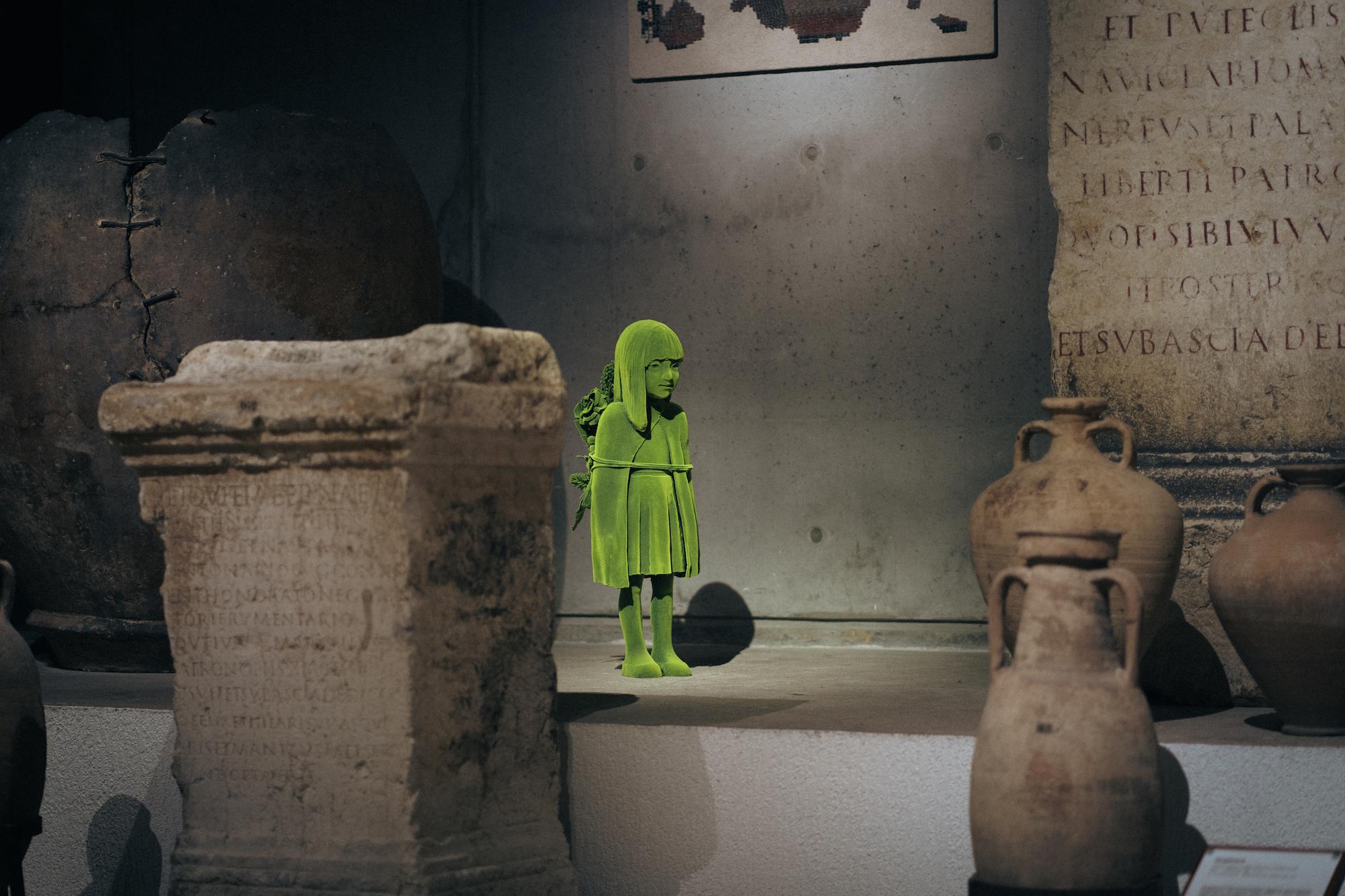 Kuvassa veistos, joka esittää vihreää sammalen peitossa olevaa tyttöhahmoa. Veistos on sijoitettu antiikinaikaisten ruukkujen ja kivipylväiden joukkoon.