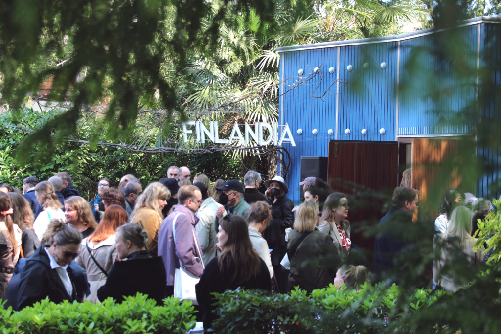 Kuva Suomen paviljongista, joka on sininen tasakattoinen rakennus vieressään iso kyltti tekstillä "Finlandia". Kymmeniä ihmisiä on kuvassa rakennuksen edessä odottamassa sisäänpääsyä ja/tai juttelemassa keskenään. Rakennusta ympäröi vehreä kasvillisuus.