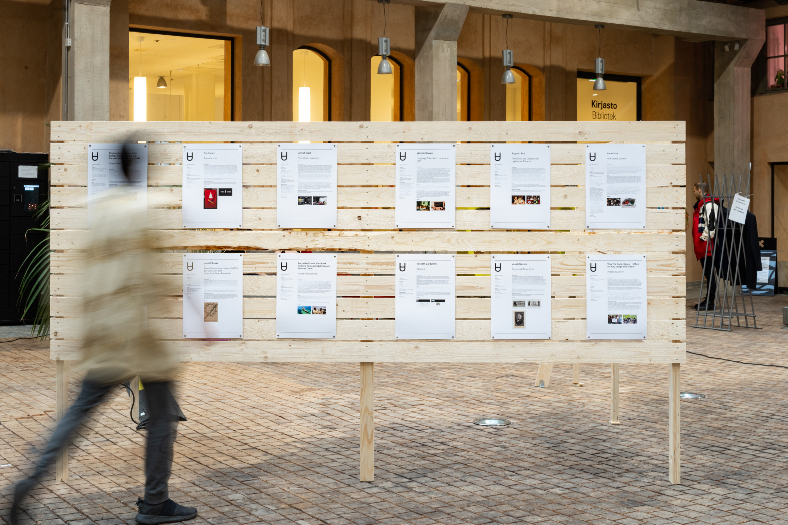 An image of a person walking and appearing blurry in front of a wooden wall-like board, which has 10 A4 size prints with images and text attached to it.
