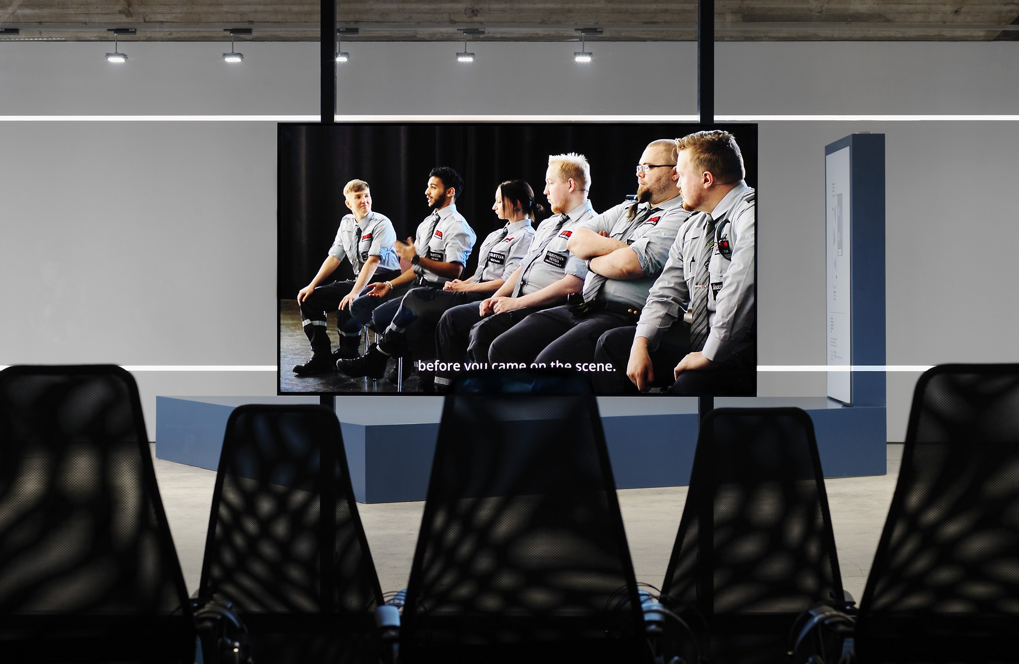 Kuva Pilvi Takalan videoinstallaatiosta Close Watch. TV-näytöllä pyörii kohtaus, jossa kuusi vartijan univormuihin pukeutunutta ihmistä istuu vierekkäin, yksi heistä puhuu ja muut kuuntelevat. Näytön edessä on viisi tuolia ja sen takana on läpinäkyvä tilanjakaja.