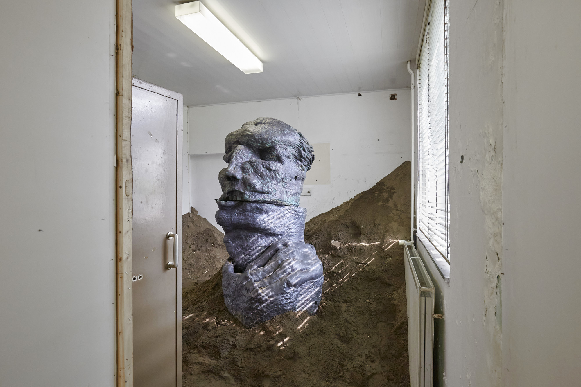 Sculpture depicting of an upper body of a person, installed in a pile of sand in a white room.