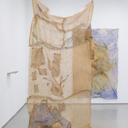 Jaana Laakkonen’s installation displayed in a gallery space with white walls. The installation consists of a textile piece hanging from the ceiling. The fabric is beige in colour and see through, probably made of jute. Another work is hanging on a wall in the background. A small shelf-looking object is attached to the wall on the left.