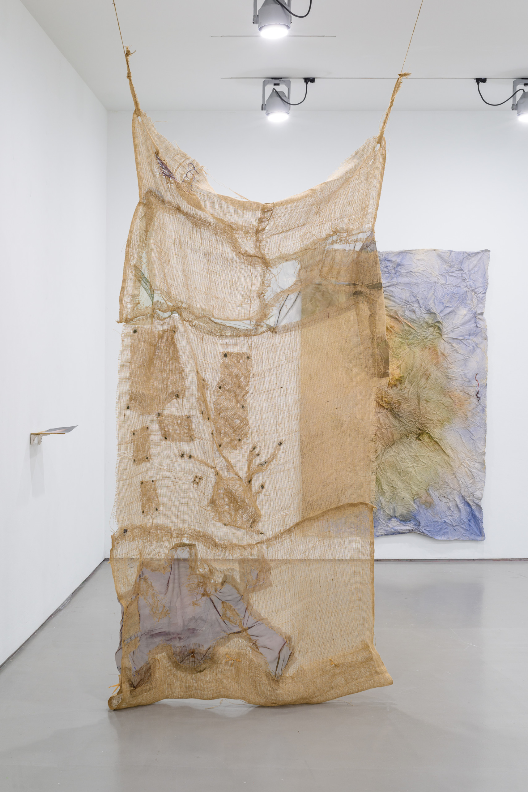 Jaana Laakkonen’s installation displayed in a gallery space with white walls. The installation consists of a textile piece hanging from the ceiling. The fabric is beige in colour and see through, probably made of jute. Another work is hanging on a wall in the background. A small shelf-looking object is attached to the wall on the left.