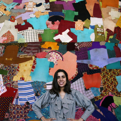 Diana Campbell Betancourt, a light brown-skinned person with shoulder-length brown hair is standing in front of a colourful backdrop comprising of a patchwork of textiles and shirts. Framed from the waist up, Diana is wearing a shirt with white and green stripes. Diana is looking into the camera and smiling while holding her hands on her waist.