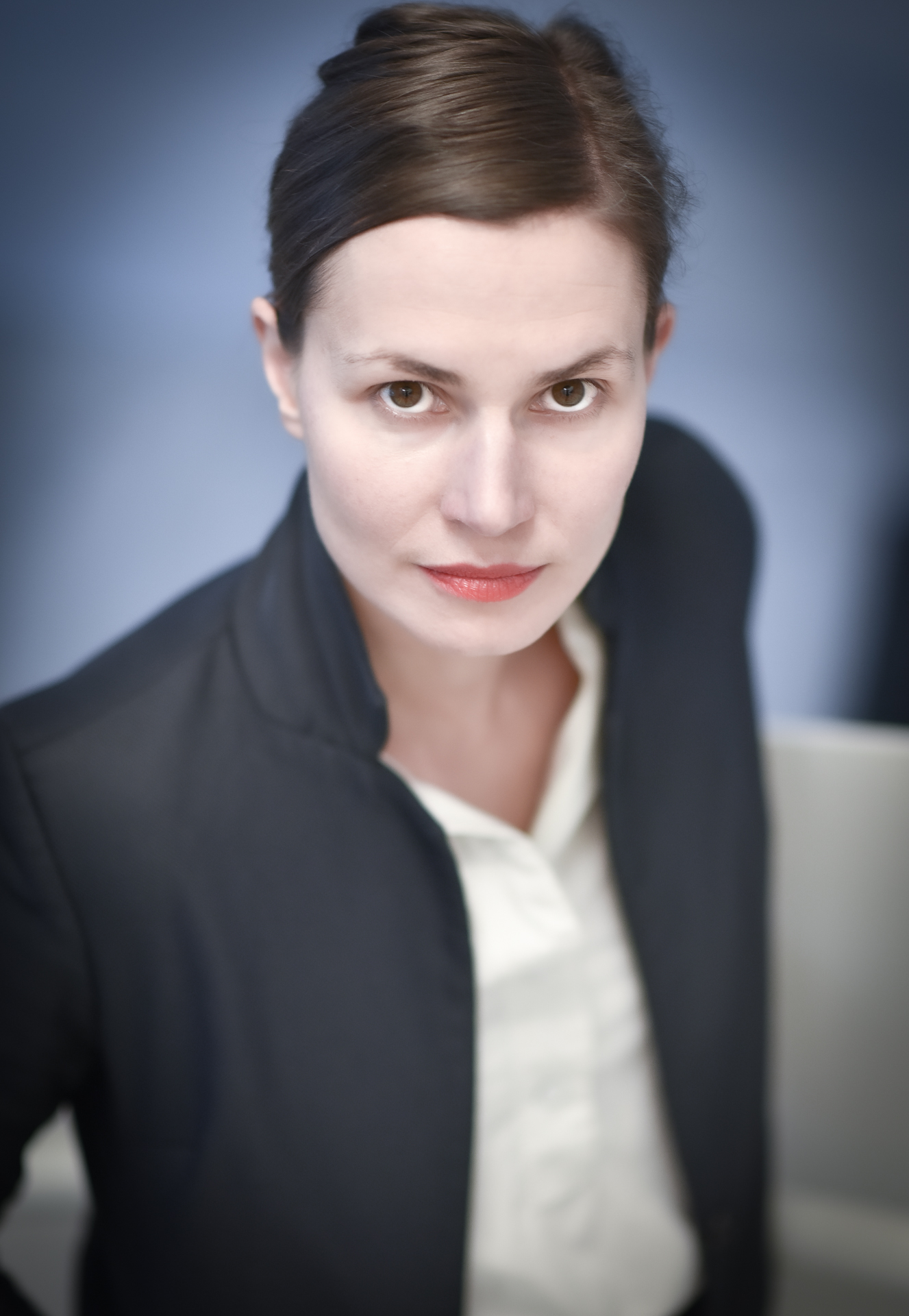 A portrait of Kasia Redzisz, a white person with brown straight hair tied back, wearing a black blazer and a white button-up shirt. The photo is taken slightly from above and she is looking directly to the camera.