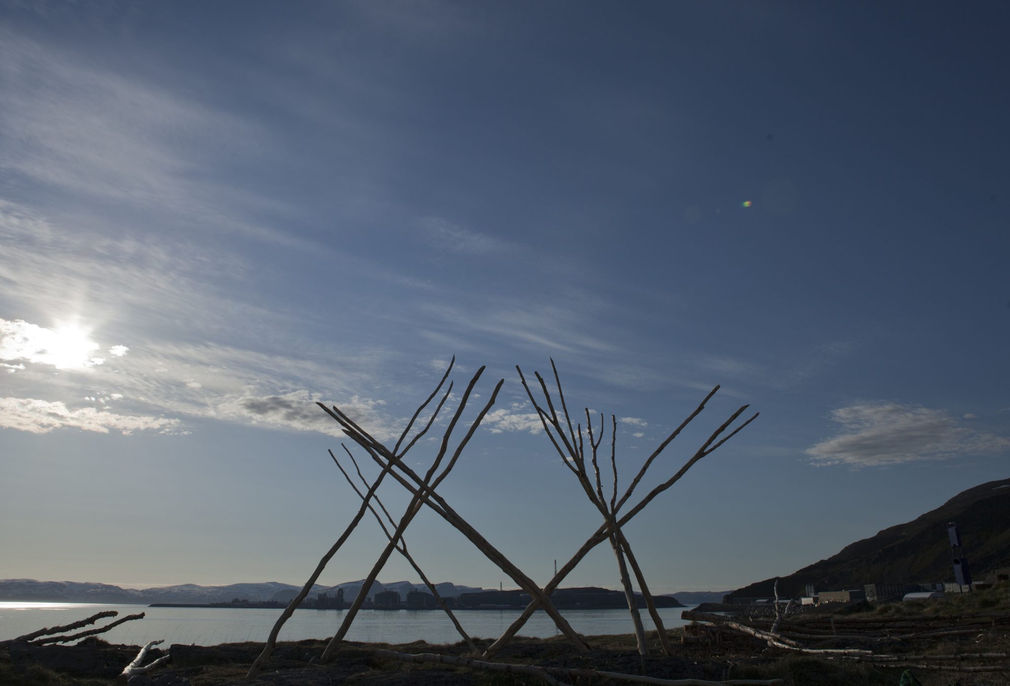 Matti Aikio's installation, Suorranaddame, features large structure comprised of wooden branches or trunks, arranged to lean on each other, forming a tent-like structure. the installation is pictured on the coast with a water body in the background.