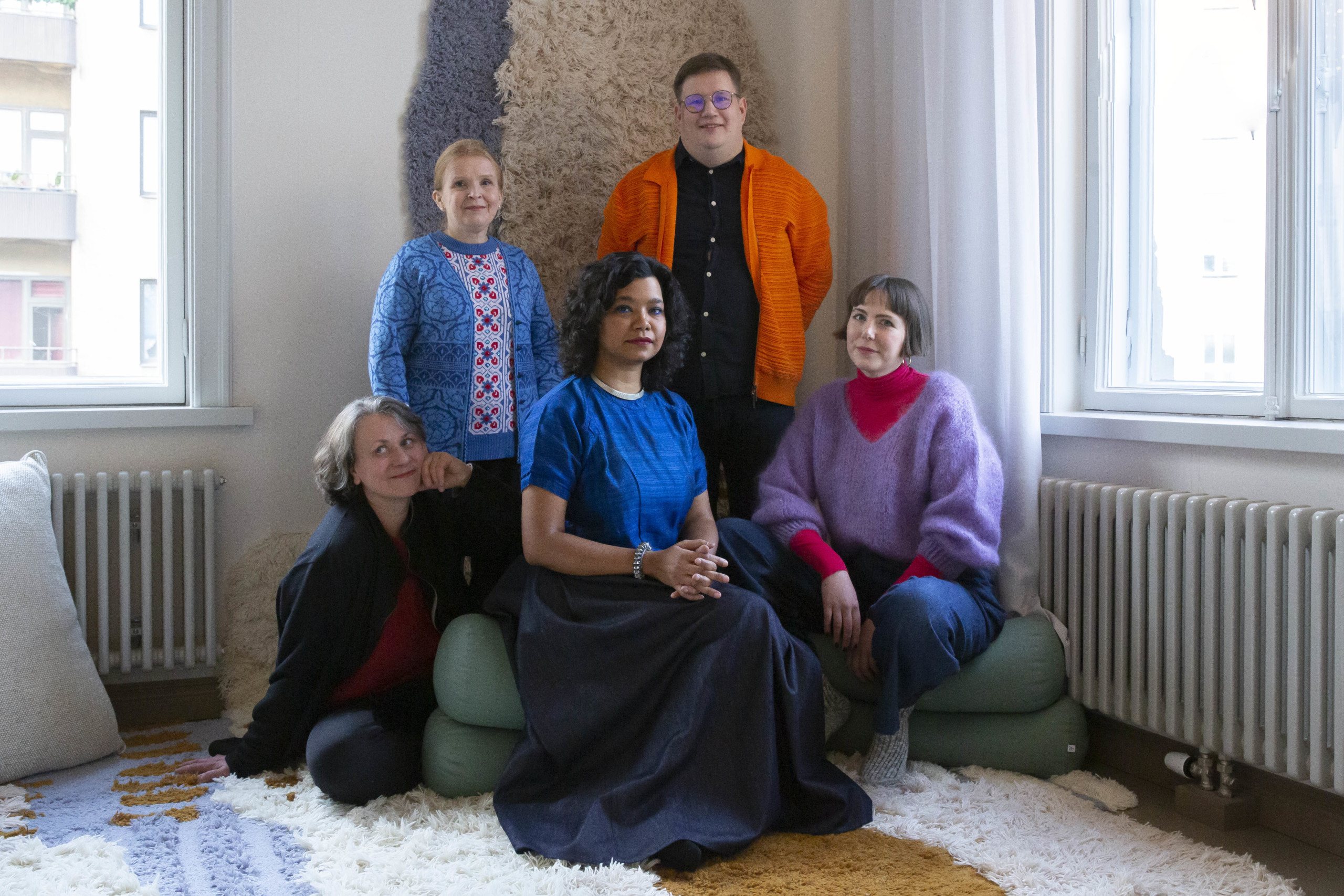 Three artists and two curators sitting next to each other and smiling at the camera. Starting from left is artist Pia Lindman, a white-skinned person with grey and brown hair, wearing a red shirt underneath a black cardigan. Next is artist Jenni-Juulia Wallinheimo-Heimonen, a white-skinned person with blonde hair, wearing a blue, white and red patterned knitted shirt and a blue cardigan on top. Next is artist Vidha Saumya, a person with light brown skin and wavy, dark brown hair and a bright blue shirt. Next to her are two curators, Jussi Koitela and . Jussi is a white-skinned with blonde hair, wearing a bright orange jacket and Yvnonne is a white-skinned person with ash-blond hair, wearing a bright lavender sweater over a pink shirt.