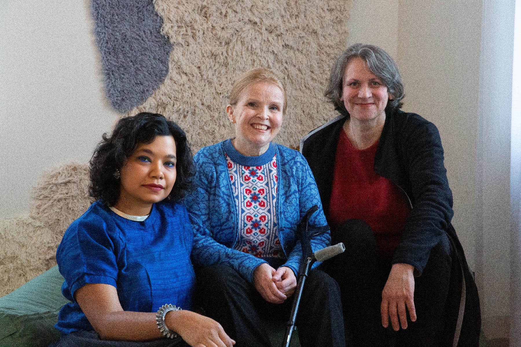 Three artists sitting next to each other and smiling at the camera. On the left is Vidha Saumya, a person with light brown skin and wavy, dark brown hair and a bright blue shirt. In the middle is Jenni-Juulia Wallinheimo-Heimonen, white-skinned with blonde hair, wearing a blue, white and red patterned knitted shirt and a blue cardigan on top. An elbow crutch is resting on her lap. On the right is Pia Lindman, with white skin and grey-brown hair, wearing a red shirt underneath a black cardigan.