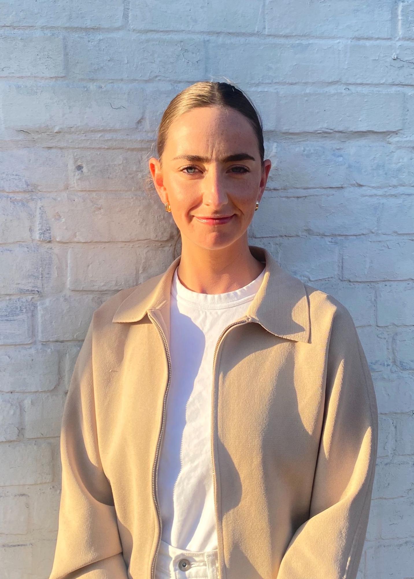 Image of Olivia Aherne, a white-skinned person photographed in sunset lighting, wearing a white t-shirt and a beige collared shirt.