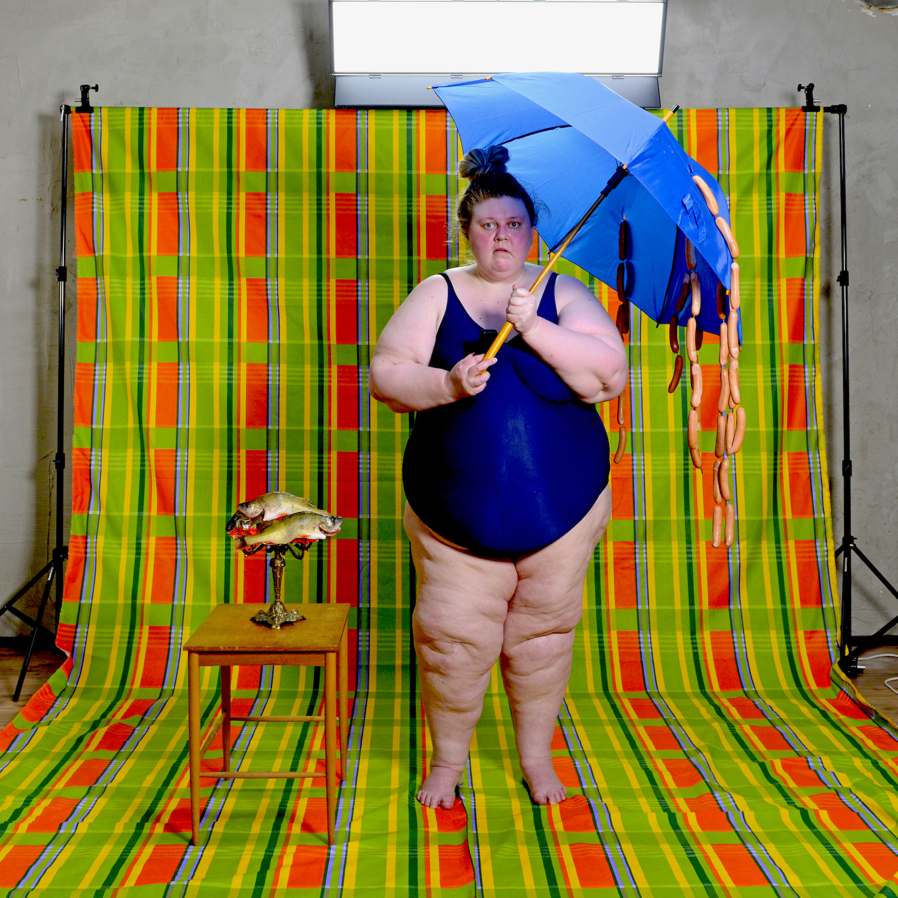 A person wearing a bathing suit in front of a colourful studio backdrop. They are holding an umbrella with sausages hanging from it. Next to them is a small table with a candle holder and toy fishes on it.