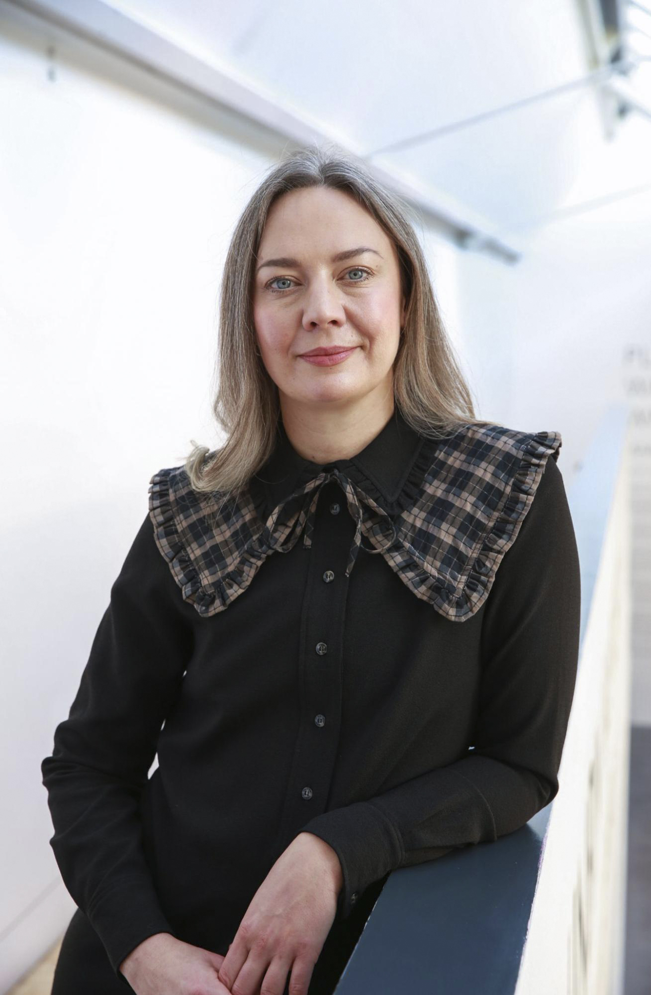 A white-skinned person with ash-blonde hair smiles into the camera. She is leaning against a staircase railing, wearing a black shirt with a large checkered collar.