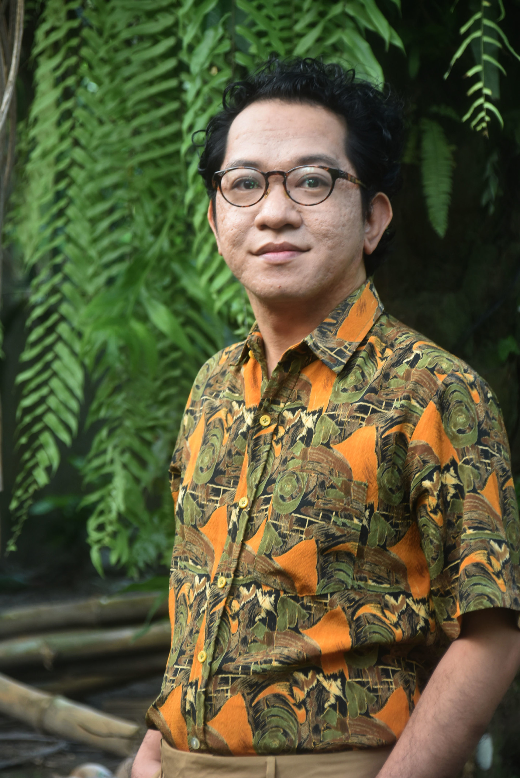 A person with short dark hair and glasses standing in front of a tree, wearing a colourful shirt.