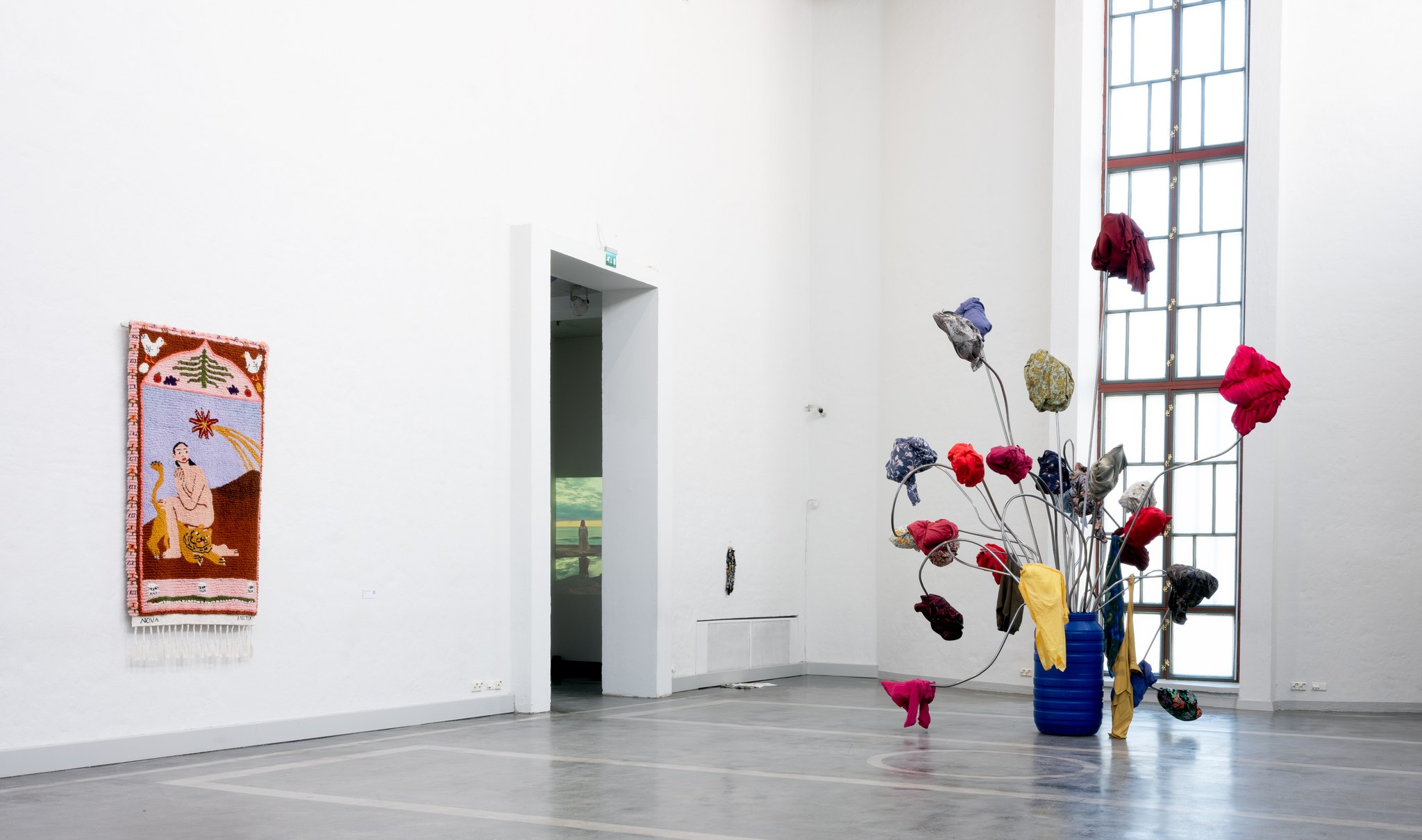 A tall exhibition space with a textile work on one of the walls and next to it a large sculpture resembling a flower pot with flowers growing out of it.