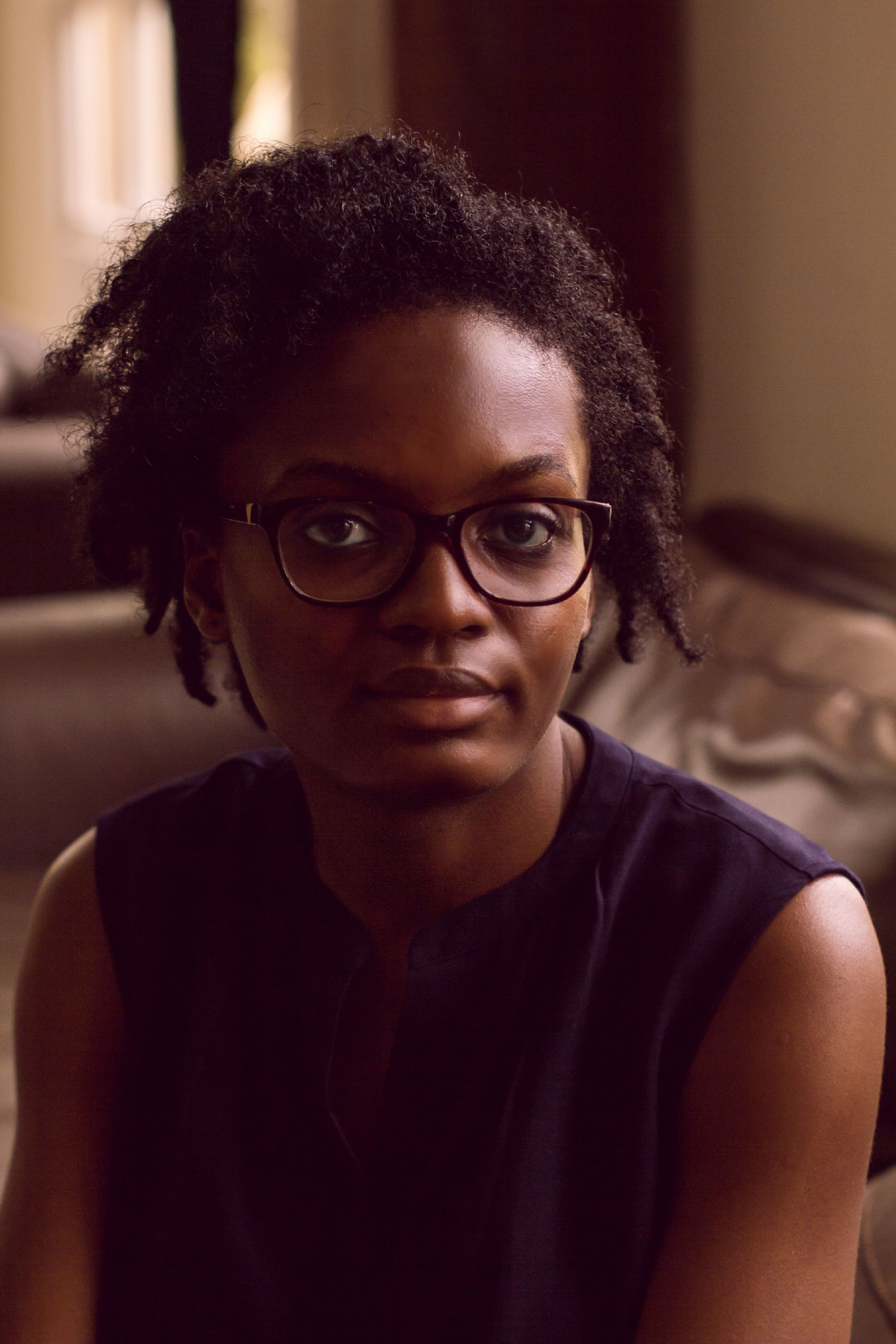 A black-skinned person with curly black hair, wearing thick black glasses and a dark sleevless top, smiles at the camera.She is pictures in an indoor space with furniture in the backgorund.