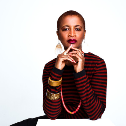 Curator Dominique Fontaine, a person with dark-brown skin, short buzz-cut black hair wearing deep pink lipstick. She is wearing a red and black striped top, ivory coloured large teardrop shaped earrings, two rings and two bangles, one made of wood and the other a stone-like material. She is looking decisively into the camera with her hands folded near her chin.