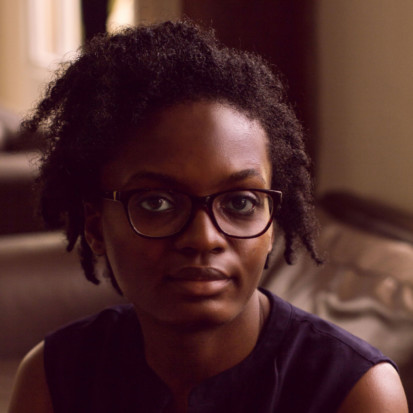 A black-skinned person with curly black hair, wearing thick black glasses and a dark sleevless top, smiles at the camera.She is pictures in an indoor space with furniture in the background.
