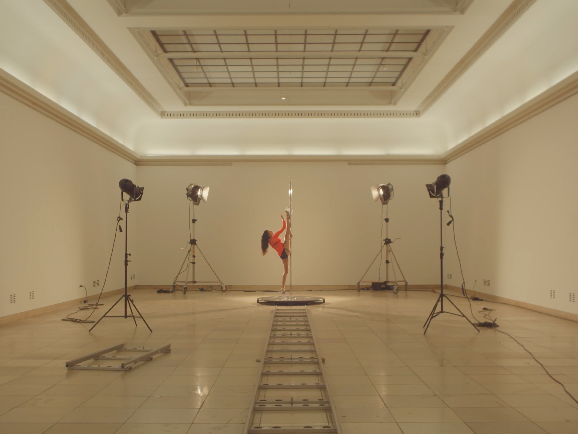 A large indoor space with four bright studio lights pointing inward at a sigular pole kept in the center. There is a person with curly blonde hair in a dance pose on the pole, with one leg and one arm vertically resting on the pole.
