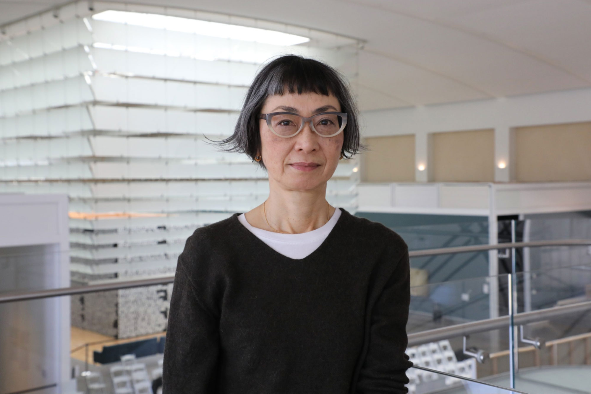 A light-skinned woman with ear-length salt and pepper hair, thick glasses wearing a black cardigan standing in a gallery.