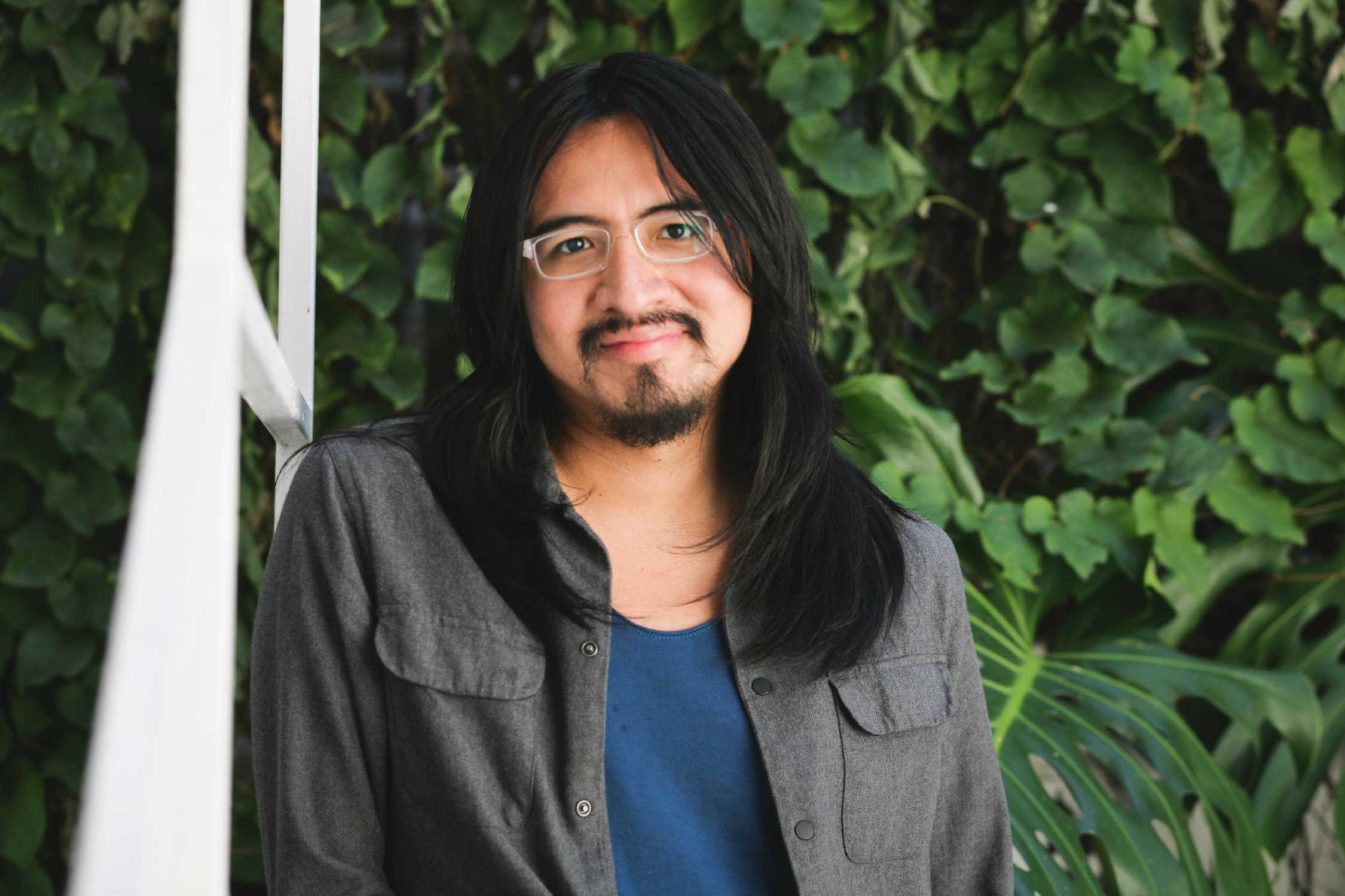 A person with light-brown skin, long straight black hair, wearing thick rectangular white frames smiles at the camera. He is wearing a grey shirt over a deep blue tshirt and is pictured against a green leafy background.
