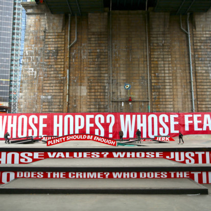 A skate park in an urban location with murals that read "Whose hopes? Whose fears? Whose justice?" among other questions. The text is written in white on red background.