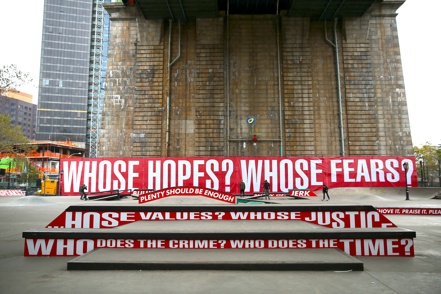 A skate park in an urban location with murals that read "Whose hopes? Whose fears? Whose justice?" among other questions. The text is written in white on red background.
