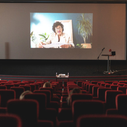 An auditorium with a large screen and rows of mostly empty red velvet seats. On the screen is a video of Ama Josephine Budge's performance. Ama, a black-skinned person with dark hair and thick framed glasses, is sitting in a room with plants and is reading from a sheet of paper.