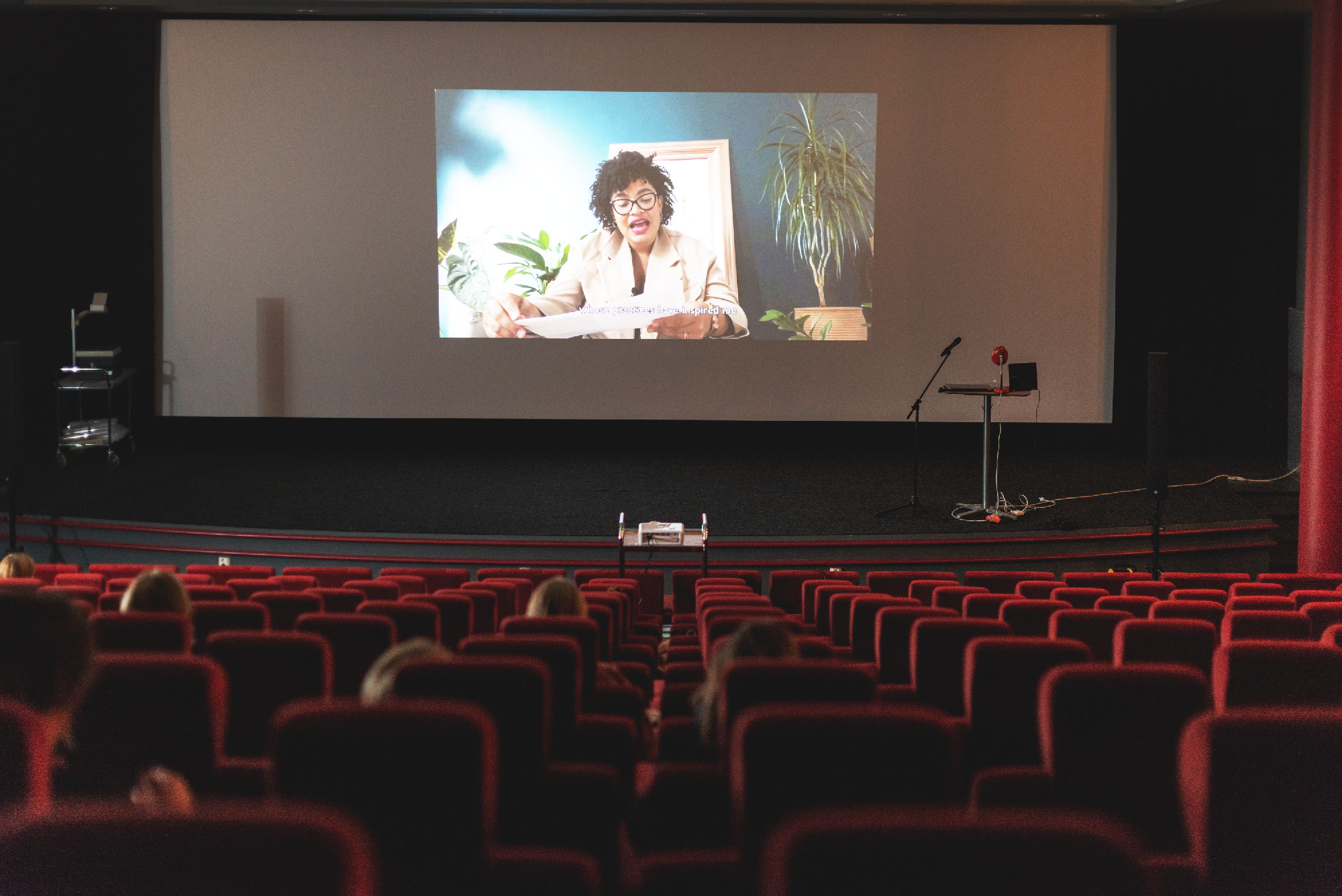 An auditorium with a large screen and rows of mostly empty red velvet seats. On the screen is a video of Ama Josephine Budge's performance. Ama, a black-skinned person with dark hair and thick framed glasses, is sitting in a room with plants and is reading from a sheet of paper.