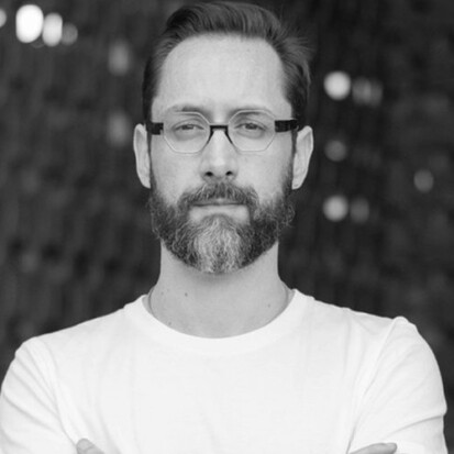A black and white photo of a light skinned person with short hair, a beard and moustache wearing glasses and a light shirt.