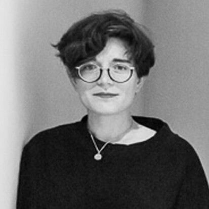 A black and white photo of a light skinned person with short dark hair wearing glasses and a dark sweater.