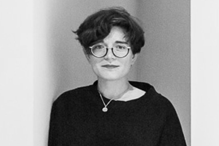 A black and white photo of a light skinned person with short dark hair wearing glasses and a dark sweater.