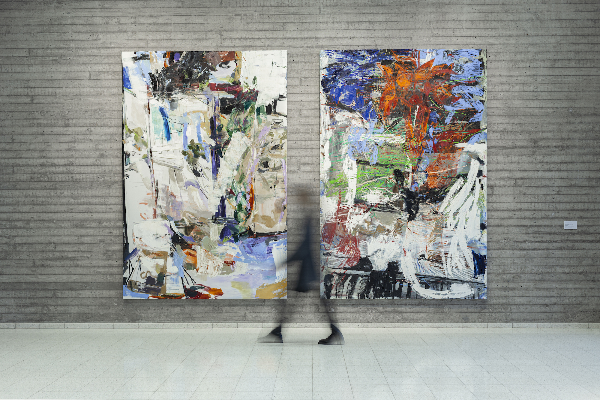 Two large colorful abstract paintings are set up against a grey wall. A blurry shape of a person is walking past the paintings, situated between them.