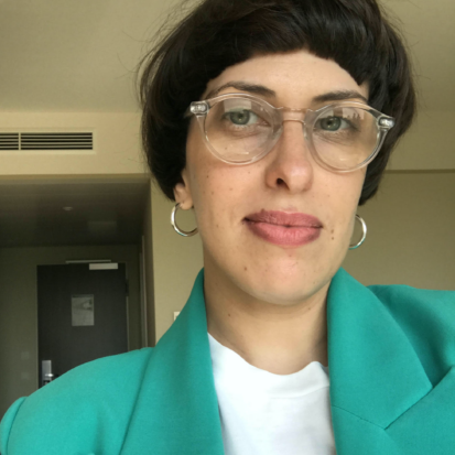 A light skinned person with short dark hair wearing glasses, a white shirt and a green jacket.