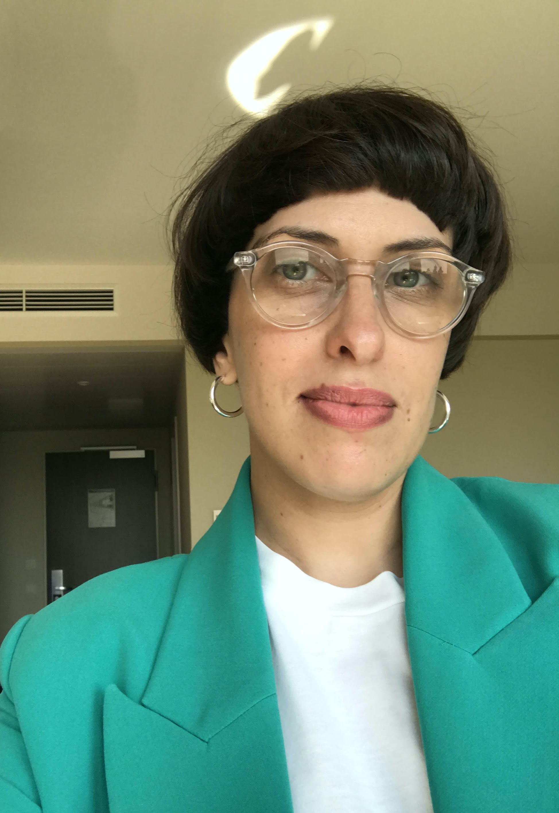 A light skinned person with short dark hair wearing glasses, a white shirt and a green jacket.
