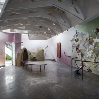 A pavilion space with colorful walls of green and red hues. On the right a sculptural textile artwork with white fabrics and colorful details. On the back a brown clay sculpture and a video screen with an abstract orange shape. A dark handrail goes through the space.