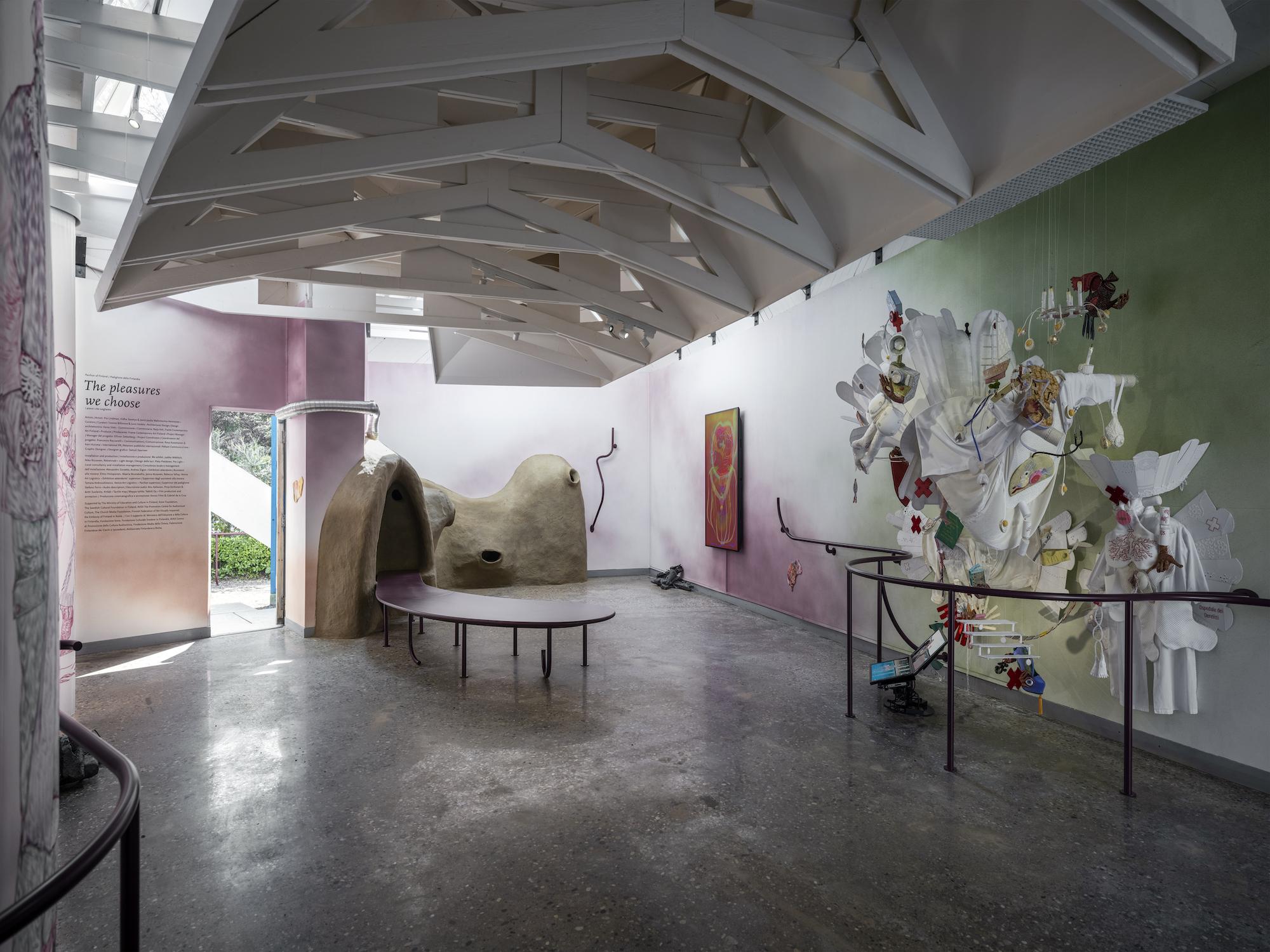 A pavilion space with colorful walls of green and red hues. On the right a sculptural textile artwork with white fabrics and colorful details. On the back a brown clay sculpture and a video screen with an abstract orange shape. A dark handrail goes through the space.